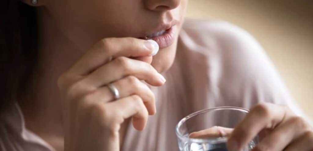 Close up young woman taking white round pill, holding water glass, unhealthy female taking painkiller to relieve headache, antidepressant or antibiotic medicine, emergency treatment