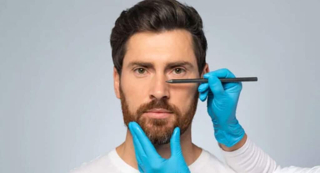 Doctor making marks on male patient's face, middle aged man on consultation at surgeon, standing on grey background
