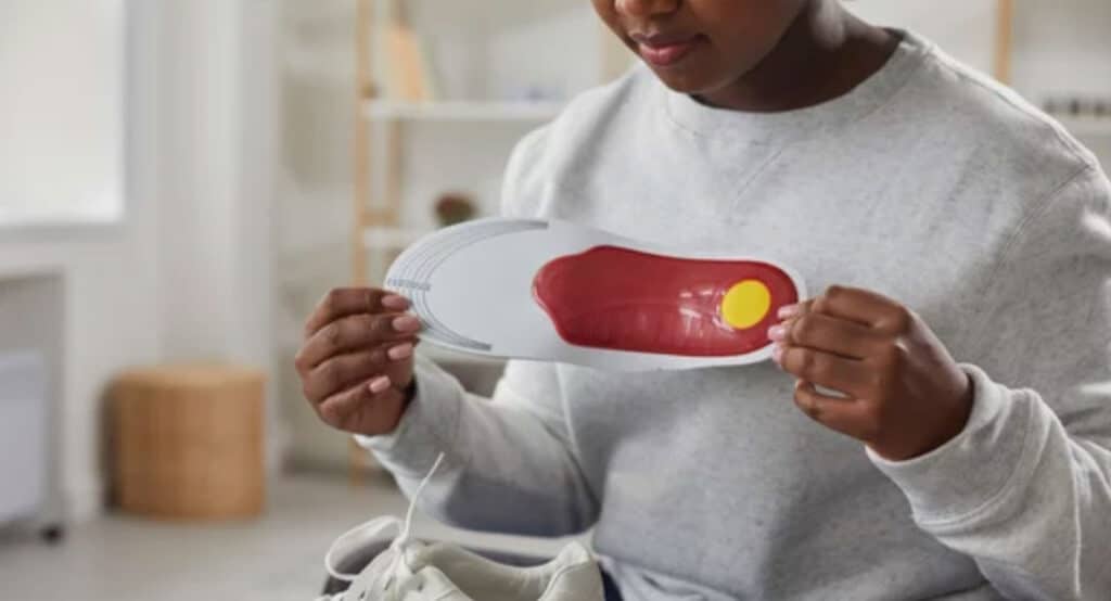 Young african american woman wearing casual gray sweater and blue jeans sitting and holding orthopedic shoe insole in her hands at home with a white sneakers on her legs.